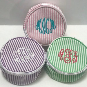 May include: Three round striped fabric zippered pouches with white piping. The pouches are in pink, purple, and green with white stripes. Each pouch has a different monogram embroidered on the front in a different color.