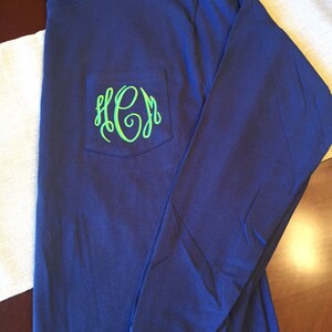 May include: A long-sleeve navy blue t-shirt featuring a chest pocket. The pocket is decorated with a bright lime green embroidered monogram consisting of the letters H, C, and M in a cursive, interlocking script font style.