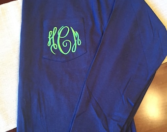 May include: A long-sleeve navy blue t-shirt featuring a chest pocket. The pocket is decorated with a bright lime green embroidered monogram consisting of the letters H, C, and M in a cursive, interlocking script font style.