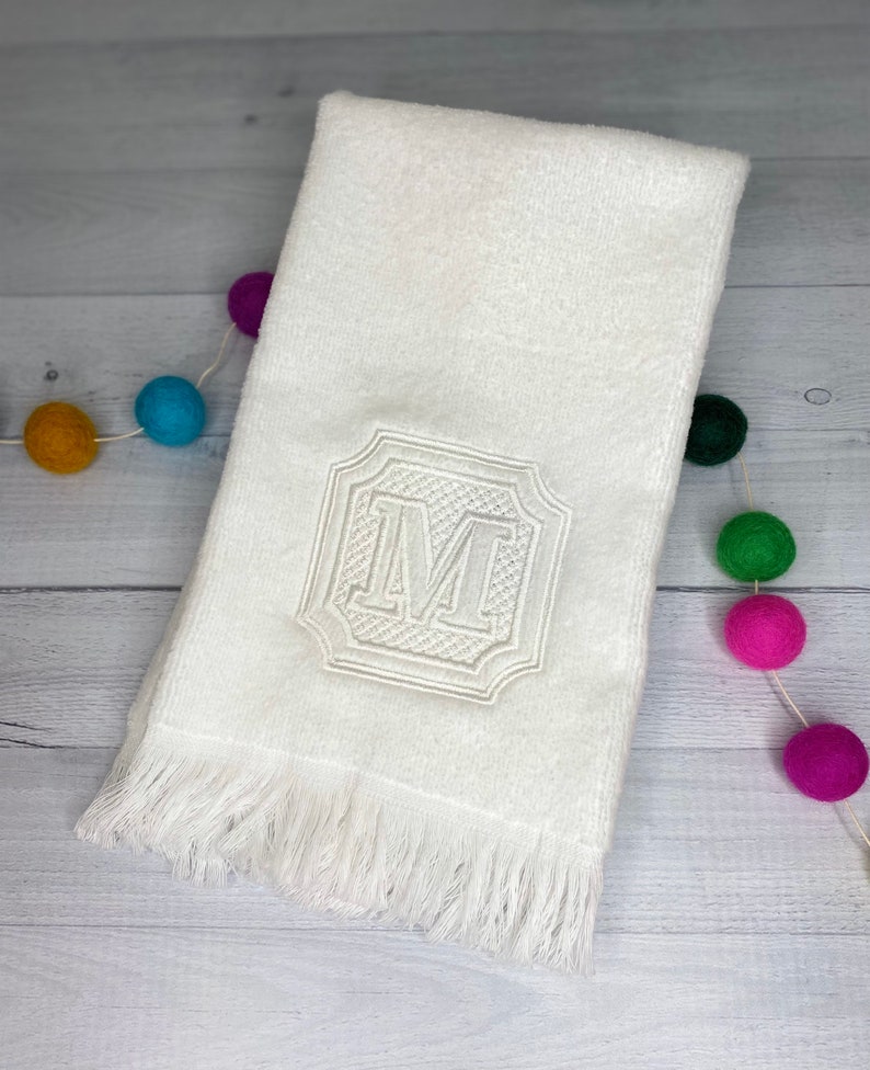 May include: White hand towel with a fringed edge and an embroidered letter "M" in a decorative frame.
