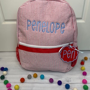 May include: Red and white striped backpack with the name Penelope embroidered in blue on the front. The backpack has a red zipper and a red tag with the word Pen on it.