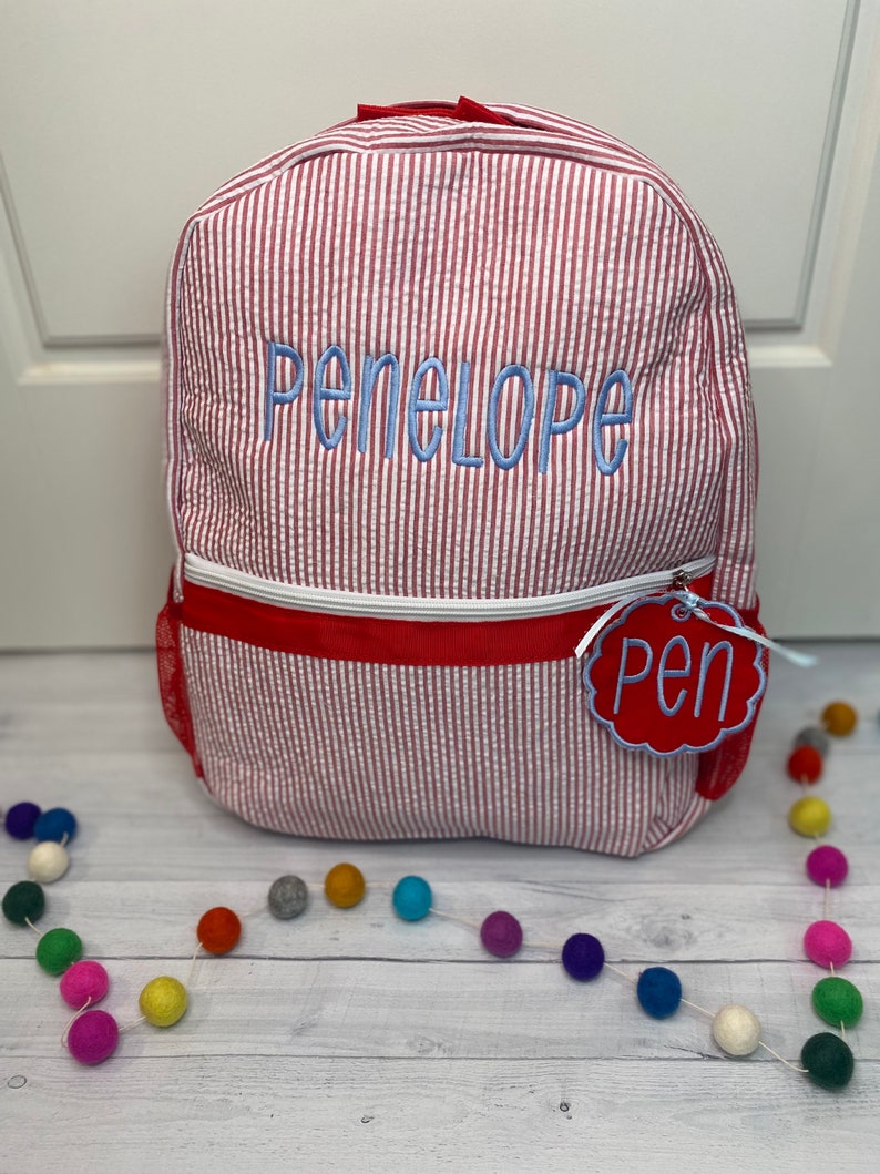 May include: Red and white striped backpack with the name Penelope embroidered in blue on the front. The backpack has a red zipper and a red tag with the word Pen on it.