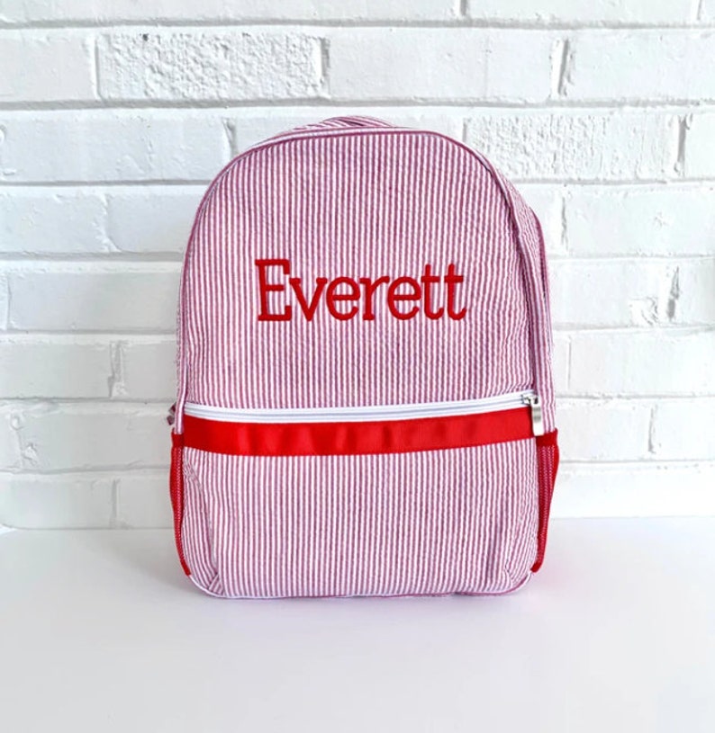 May include: Pink and white striped backpack with a red zipper and red trim. The backpack is personalized with the name "Everett" in red embroidery.