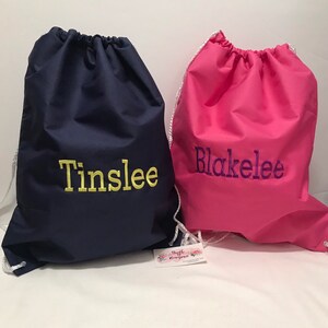 May include: Two personalized drawstring bags, one navy blue with the name "Tinslee" embroidered in yellow, and one pink with the name "Blakelee" embroidered in purple.