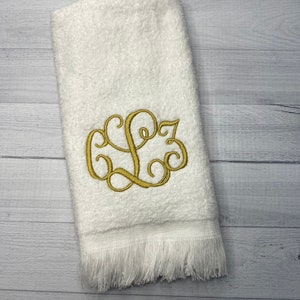 May include: White hand towel with a gold embroidered monogram of the letters "L.S." in a script font. The towel has a white fringe border.