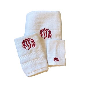 May include: A set of three white terry cloth towels featuring a red embroidered monogram. The towels include one large bath towel, one hand towel, and one washcloth, each displaying a decorative script letter design in the center of the fabric.