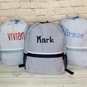 May include: Three blue and white striped backpacks with personalized names: Vivian, Mark, and Grace. The backpacks have a navy blue accent and a zipper closure.