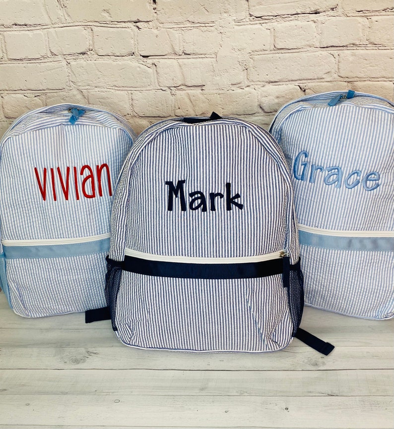 May include: Three blue and white striped backpacks with personalized names: Vivian, Mark, and Grace. The backpacks have a navy blue accent and a zipper closure.