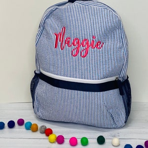 May include: A blue and white striped backpack with a pink embroidered name "Maggie" on the front. The backpack has a navy blue zippered pocket and mesh side pockets.