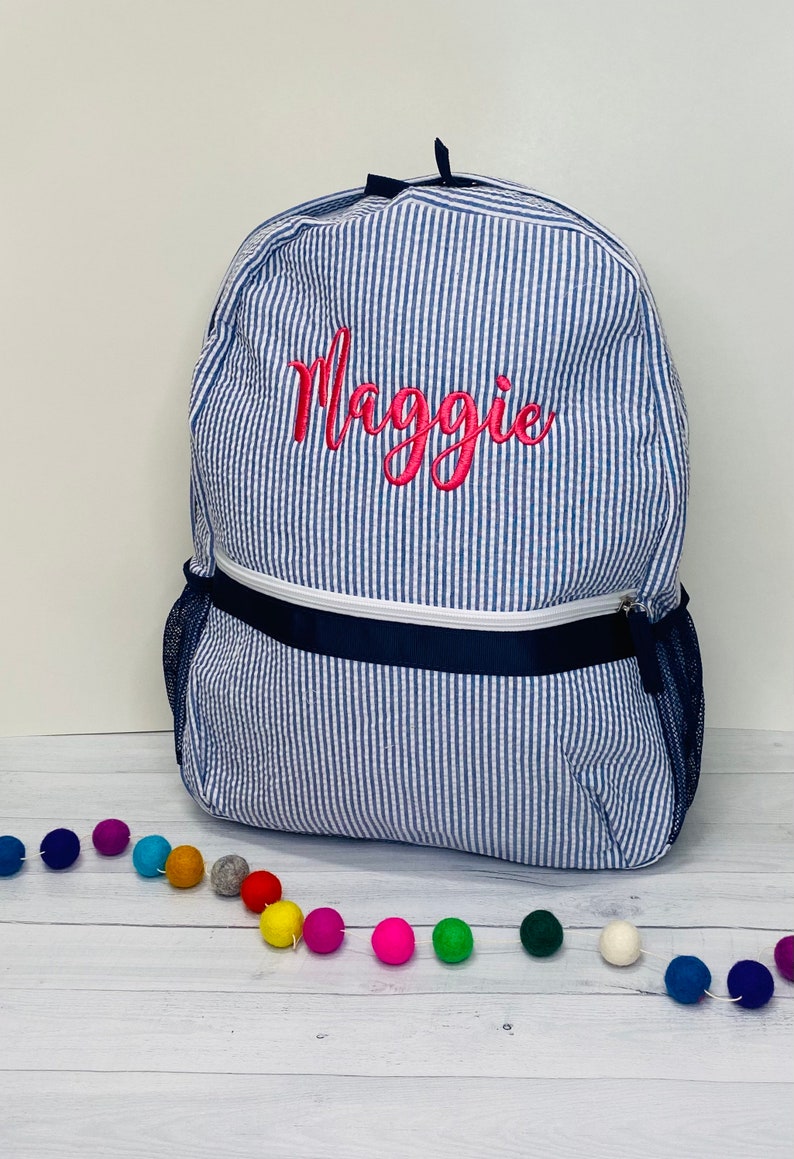 May include: A blue and white striped backpack with a pink embroidered name "Maggie" on the front. The backpack has a navy blue zippered pocket and mesh side pockets.