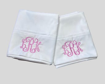 May include: Two white pillowcases featuring a decorative pink embroidered monogram with the letters LRK in a cursive script style. The fabric is smooth and opaque, with a horizontal stitched hem detail visible across the top of each pillowcase.