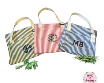 May include: Three rectangular fabric bags with vertical stripes in olive green, coral red, and blue. Each bag features a light-colored shoulder strap with a black buckle and a personalized monogram embroidered on the front in a contrasting thread color.