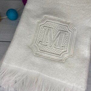 May include: White hand towel with a white embroidered letter "M" in a decorative frame. The towel has a white fringe border.