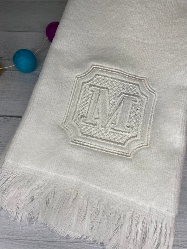 May include: White hand towel with a white embroidered letter "M" in a decorative frame. The towel has a white fringe border.