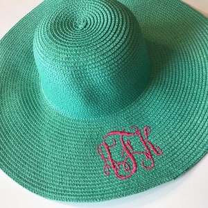 K&ouml;nnte beinhalten: Ein blaugr&uuml;ner Sonnenhut mit einem rosa gestickten Monogramm. Der Hut ist aus gewebtem Stroh.