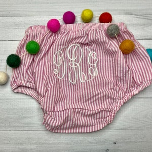 May include: Pink and white striped baby bloomers with a white embroidered monogram. The bloomers are on a white wooden surface with a colorful garland of pom-poms.