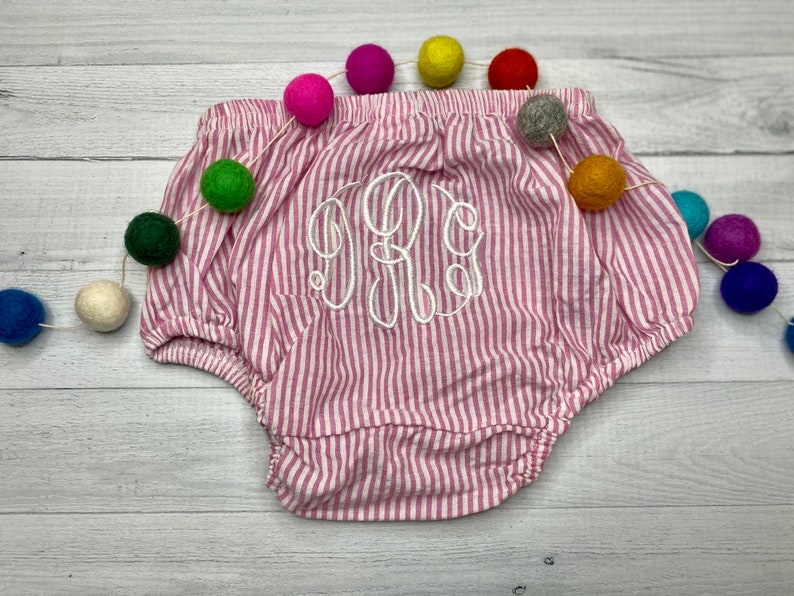May include: Pink and white striped baby bloomers with a white embroidered monogram. The bloomers are on a white wooden surface with a colorful garland of pom-poms.