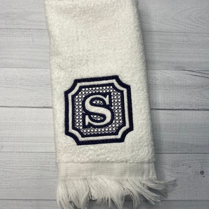 May include: White hand towel with a navy blue embroidered monogram of the letter "S" in a decorative frame.