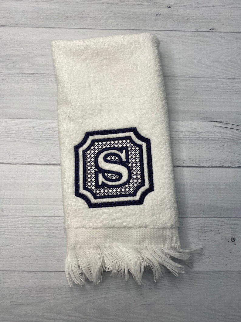 May include: White hand towel with a navy blue embroidered monogram of the letter "S" in a decorative frame.