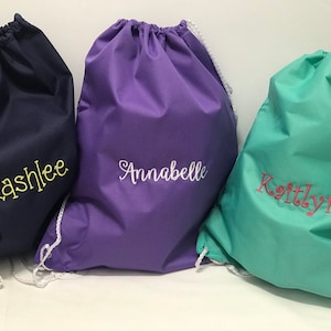 May include: Three drawstring bags in different colors with personalized names. The bag on the left is navy blue with the name "Kashlee" embroidered in yellow. The middle bag is purple with the name "Annabelle" embroidered in white. The bag on the right is teal with the name "Kaitlyn" embroidered in pink.