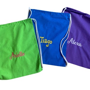 May include: Three drawstring bags with different colored fabric and personalized names. The green bag says "Arielle" in pink, the blue bag says "Tiago" in yellow, and the purple bag says "Alexa" in white.