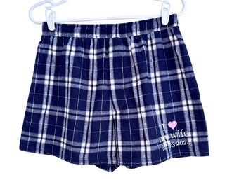 Può includere: Pantaloncini a quadri blu navy e bianchi con elastico in vita su una gruccia di plastica bianca. In basso a destra è presente una piccola icona a forma di cuore rosa sopra il testo bianco che recita I love my wife Est. 2024.