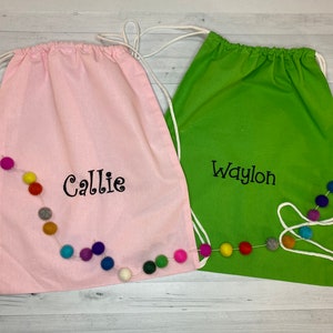 May include: Two personalized drawstring bags with pom pom trim. The pink bag says "Callie" and the green bag says "Waylon".
