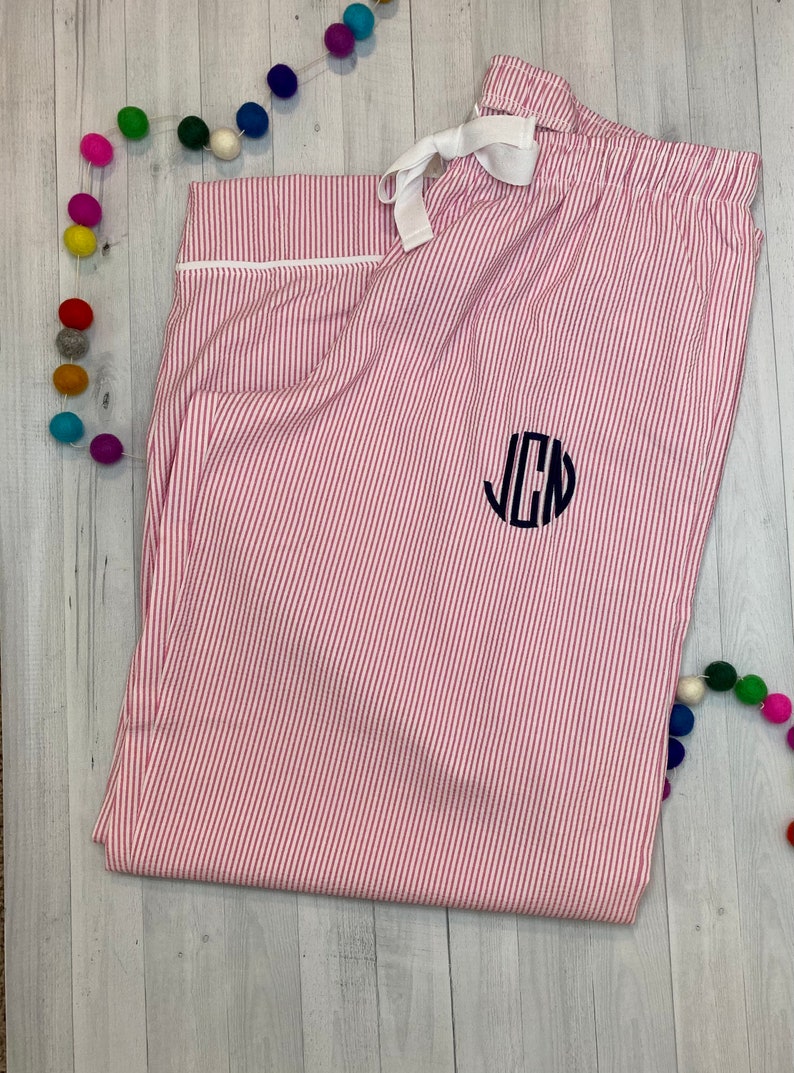 K&ouml;nnte beinhalten: Rosa-wei&szlig;e gestreifte Schlafanzughose mit wei&szlig;em Kordelzug und einem schwarzen Monogramm der Buchstaben "JCN" auf der Vorderseite.