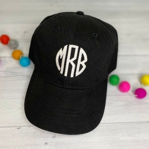 May include: Black baseball cap with a white embroidered monogram of the letters MRB in a circular design. The cap has a curved brim and a button on top. A string of colorful pom-poms decorates the background.