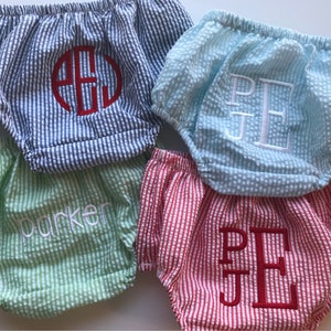 May include: Four pairs of baby bloomers in different colors and patterns. The bloomers are made of a soft, lightweight fabric and have a ruffled elastic waistband. The bloomers are embroidered with different names, including "Parker" and "P.E.J."