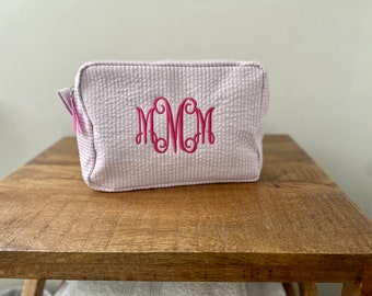 May include: A rectangular zippered pouch made of light pink and white seersucker fabric. The front of the pouch features a large, embroidered monogram in bright pink thread with the letters M, M, and M in a decorative script font.