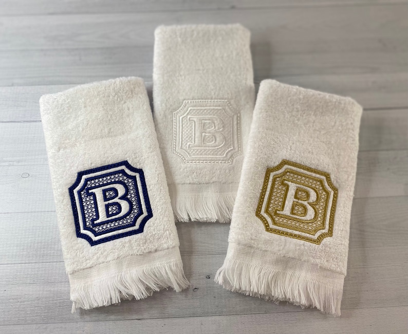 May include: Three white towels with fringed edges, each embroidered with a different colored letter "B" in a decorative frame. The towels are arranged on a light wood surface.