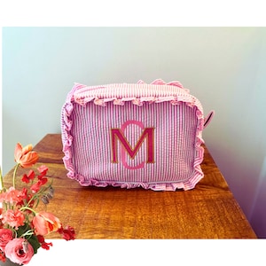 May include: A pink and white striped makeup bag with a ruffled edge and a pink and gold monogram "M".