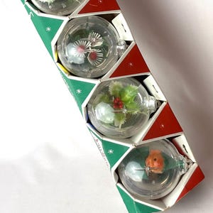 May include: A rectangular cardboard box with a red and green geometric pattern containing four clear spherical ornaments. Each ornament holds a small decorative element inside, including holly leaves with red berries and a small orange figurine.
