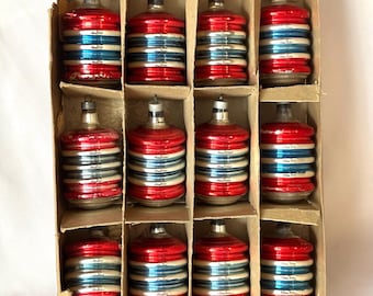 May include: A set of twelve cylindrical glass ornaments arranged in a cardboard box. Each ornament features alternating horizontal bands of metallic red, silver, and blue, topped with a silver-colored metal cap and a small hanging loop.