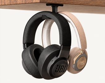 May include: A black under-desk headphone hanger mounted to a wooden surface, holding two pairs of over-ear headphones. One pair is black with the JBL logo, and the other pair is beige and metallic gold with the B&O logo on the ear cup.