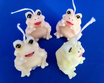 May include: Four small frog-shaped candles, two are white and two are a pale yellow. The candles have white wicks and painted details, including eyes, a mouth, and legs.