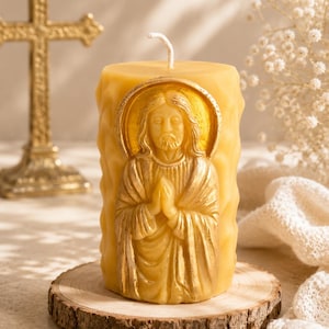 Puede incluir: Una vela cilíndrica de cera de abeja amarilla con un relieve dorado de una figura con cabello largo y manos juntas en oración dentro de un halo, con una superficie texturizada ondulada y una mecha blanca individual.