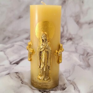 Puede incluir: Una vela de pilar color crema con una figura religiosa dorada, un halo y dos cruces. La vela tiene una superficie lisa y una mecha. La figura dorada está en posición de oración. Objeto religioso.