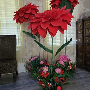 May include: A display of large, red paper flowers with green stems and leaves. The flowers are in a wooden planter with smaller, colourful flowers and greenery. The arrangement is set against a neutral background.