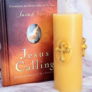 Puede incluir: Una vela amarilla con un diseño de cruz dorada. La vela está junto a un libro titulado "Jesus Calling: Enjoying Peace in His Presence" de Sarah Young.