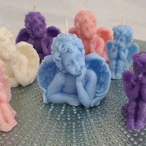 Puede incluir: Una colección de velas con forma de querubín en varios colores pastel, incluyendo rosa, azul, morado y crema. Cada vela presenta alas detalladas y una expresión serena. Las velas están dispuestas sobre una superficie texturizada de color plateado.