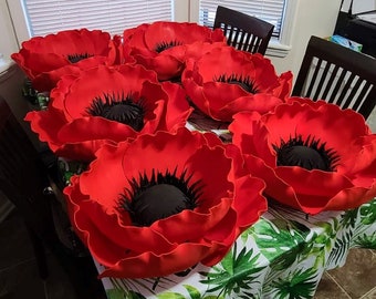 May include: A table covered in a green and white floral tablecloth with eight large red paper flowers. The flowers have black centers and are arranged in a circle.