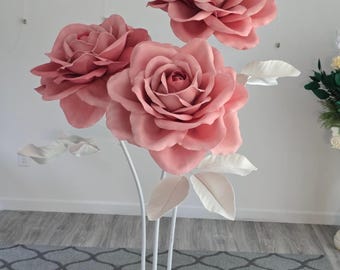 May include: Three large, pink paper roses with white leaves and stems. The flowers are mounted on a white metal stand. The roses are a dusty rose color and are arranged in a decorative display.