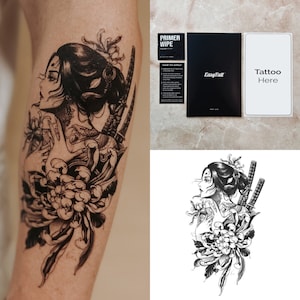 Puede incluir: Tatuaje temporal en blanco y negro de una mujer con el pelo largo, sosteniendo dos espadas, con una flor de crisantemo detrás de ella. El tatuaje está en el brazo de una persona. La imagen también incluye una hoja de instrucciones para aplicar el tatuaje.
