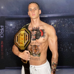 May include: A man wearing white shorts holds a gold and black UFC championship belt. He has several tattoos on his chest and arm, including a tiger, a gorilla, and the words "Gregor" and "Gorious".