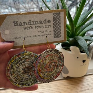 May include: Handmade paper earrings with a spiral design in shades of red, yellow, and green. The earrings are held in front of a tag that reads "Handmade with love by: Izabella". A small succulent plant in a white dog-shaped pot is in the background.