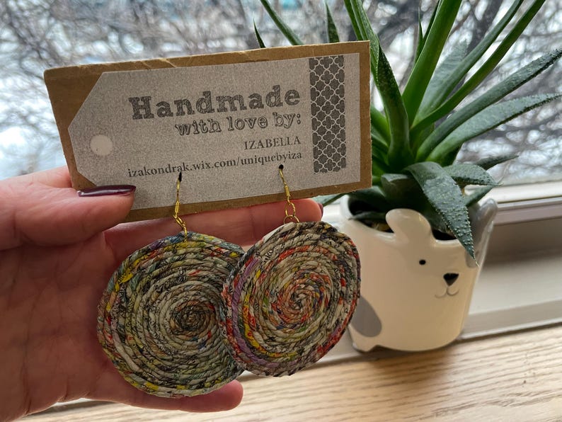 May include: Handmade paper earrings with a spiral design in shades of red, yellow, and green. The earrings are held in front of a tag that reads "Handmade with love by: Izabella". A small succulent plant in a white dog-shaped pot is in the background.