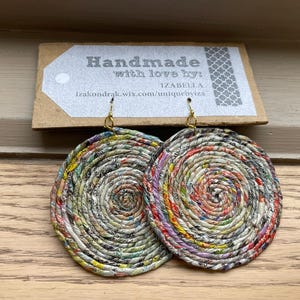 Puede incluir: Pendientes circulares de papel hechos a mano con un diseño en espiral. Los pendientes tienen ganchos dorados. El papel es multicolor, con tonos rojos, amarillos, verdes y azules. La etiqueta sobre los pendientes dice "Handmade with love by: IZABELLA".