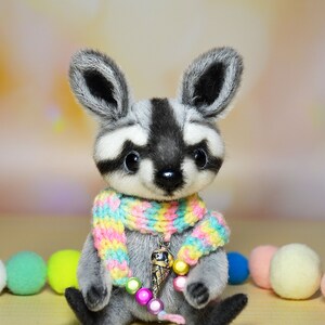 May include: A handmade plush raccoon with a gray and white face, black eyes, and large ears. It wears a colorful scarf and a charm necklace with an ice cream cone pendant. The raccoon is sitting with colorful pom-poms in the background.
